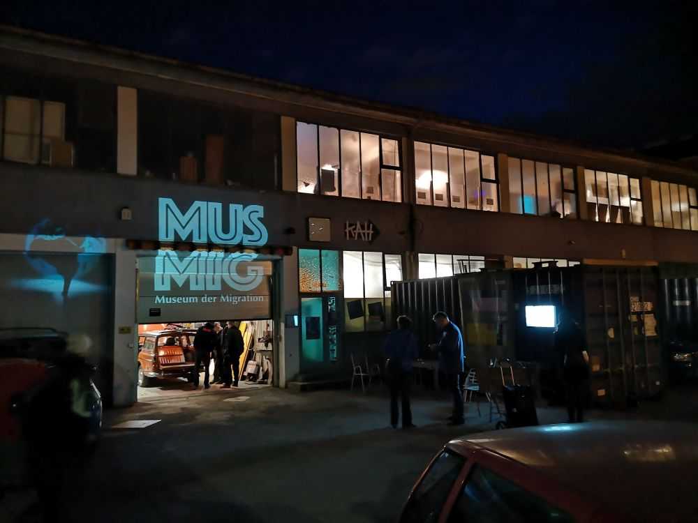 WIENWOCHE 2023: MUSMIG - Das Plenum. A night image of a two stores building. A garage door open, where a projection of the logo MUSMIG is visible. © MUSMIG (LjB)